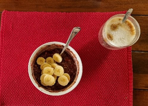 Ein rotes Stoff-Tischset auf einem Holztisch. Auf dem Set ein Glasbecher mit Cappuccino, in der Schale dunkles Porridge mit Bananenscheiben.