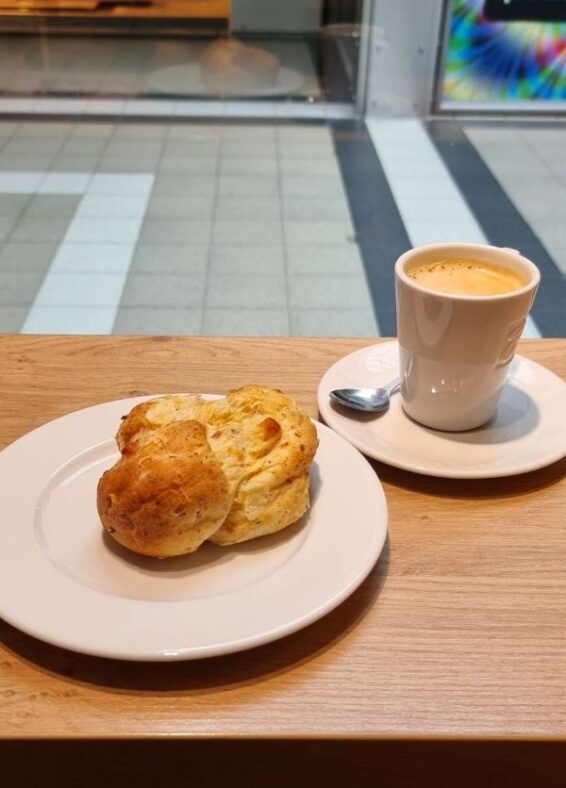 Ein Teller mit einem Brioche, daneben eine Tasse Kaffee. Beides steht auf einem Holztisch mit Blick in einen Gang im Einkaufszentrum.