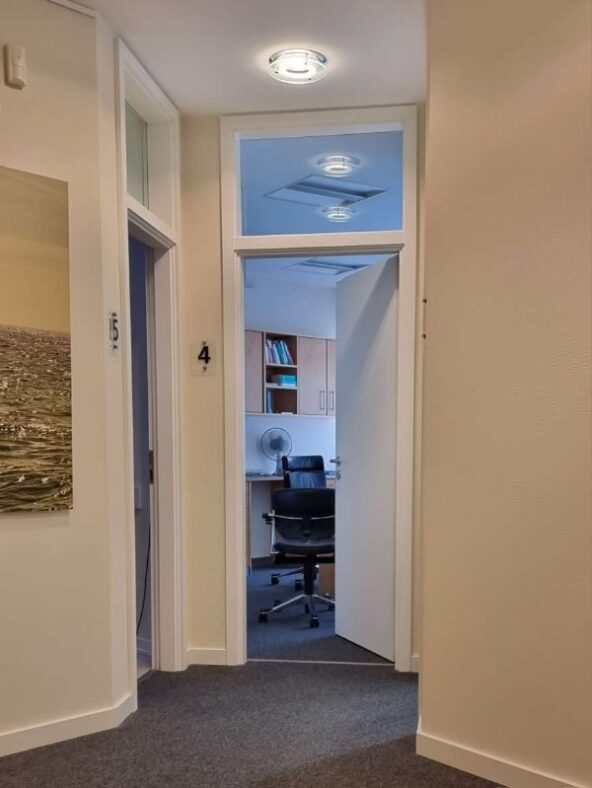 Im Flur der Arztpraxis geht der Blick auf die geöffnete Tür eines Behandlungsraums mit Schreibtisch und Büchern.