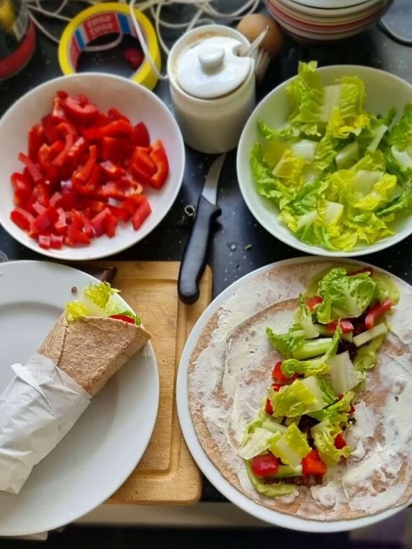 Auf dem Bild Zutaten für Tortilla-Wraps, einer ist schon gerollt. Belegt sind sie mit Frischkäse, Guacamole, roter Paprika, Salatgurke und Romana-Salat. Im Hintergrund Kabel und Werkzeuge.