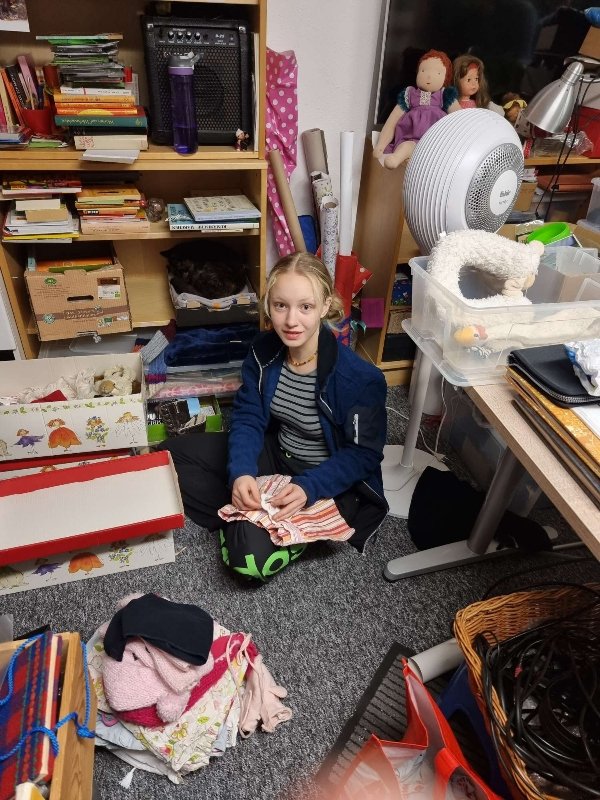 Die Tochter der Autorin sitzt auf dem Boden im Keller, umgeben von Büchern, Puppen, Kleidungsstücken, die sie gerade aussortiert.