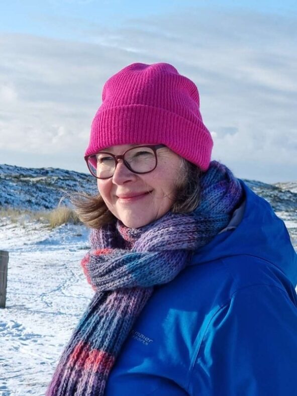 Die Autorin in bunter Winterkleidung, zufrieden lächelnd vor verschneiten Dünen. Das Lächeln zeigt ihre Freude an der Entschleunigung.