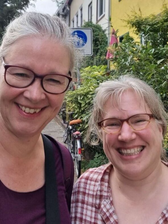 Zwei Frauen sehr unterschiedlicher Körpergröße lachen in die Kamera, dahinter eine kleine Gasse mit viel Straßengrün