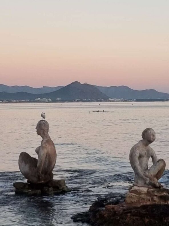 Tonfiguren in Strandnähe sitzen im Meer in Can Picafort auf Mallorca, dahinter rosa Morgenhimmel