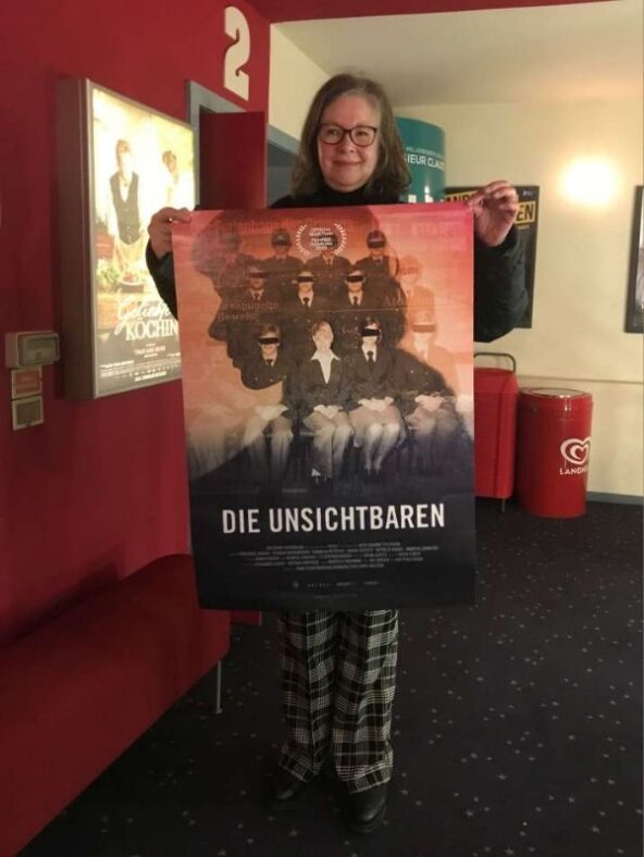 Die Autorin hält das Filmplakat des Films "Die Unsichtbaren" und steht im Foyer des Blankeneser Kinos vor dem Sall 2.