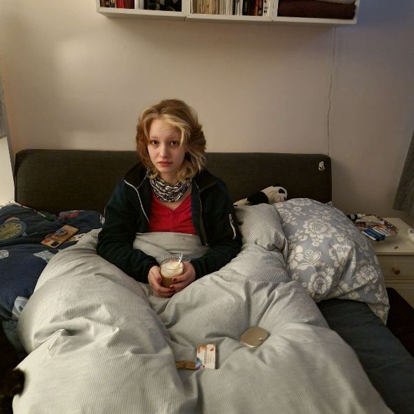 Eine blonde junge Frau sitzt blass und erkältet in einem Bett mit grauem Stoffrücken und heller Bettwäsche.