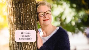 Die Autorin lehnt verträumt an einem Baumstamm, auf dem ein Schild klebt mit der Aufschrift: 44+ Tipps für mehr Körperliebe