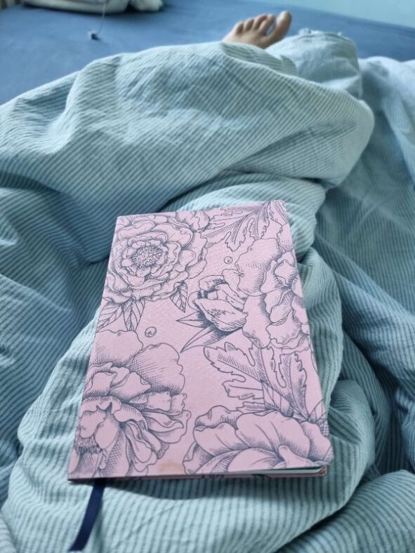 Ein Notizbuch im rosa Einband mit floralen Ornamenten in Blaugrün liegt auf einer grünlichen Bettdecke.