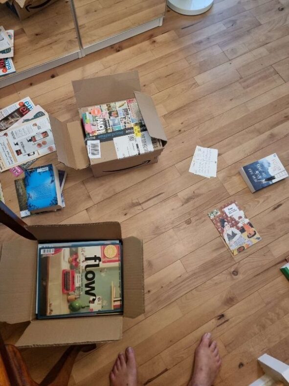 Auf hölzernem Fußboden liegen allerlei Zeitschriften, Bücher und zwei Pakete.