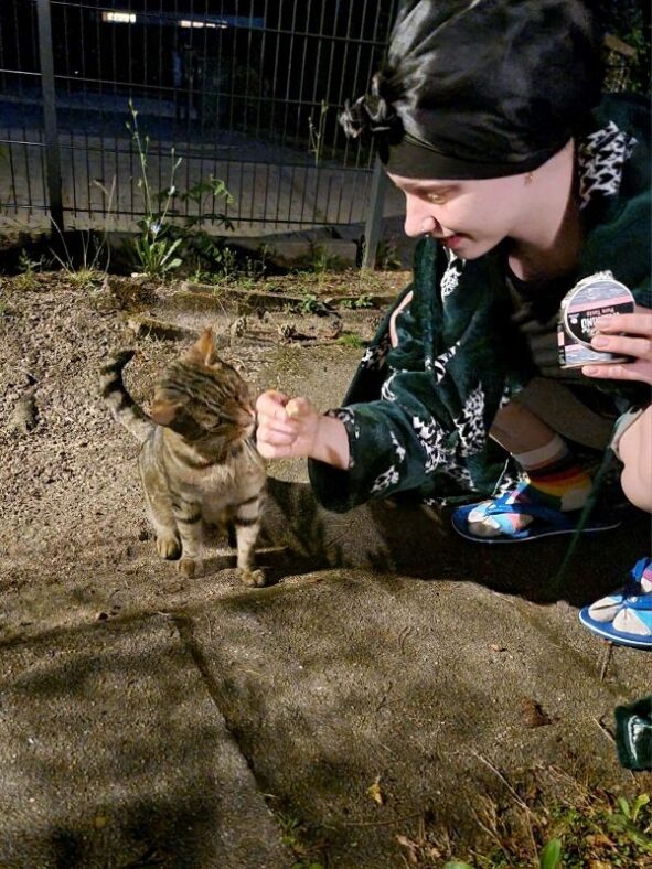 Dit Tochter der Autorin miteiner schwarzen Haarhaube auf dem Kopf und im grünen Hausmantel mit Wildkatzenmotiv hockt am Boden, mit einer Dose in der Hand, und streichelt eine getigerte Katze.