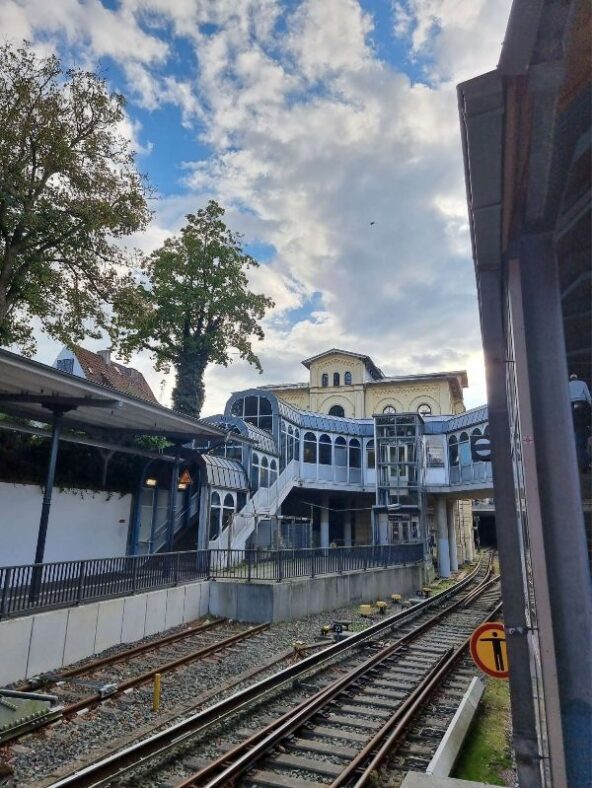 Ein Panoramabild vom Blankeneser Bahnhofsgebäude, vom Gleis aus fotografiert. Zu sehen ist der Abgang zum Nachbargleis mit interessanter Architektur, die Brücke über die Gleise, und darüber ein wolkig-blauer Vorabendhimmel.