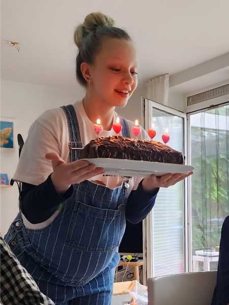Die Tochter der Autorin steht hochschwanger in einer gestreiften Latzhose am Tisch, strahlend mit einem Geburtstagskuchen mit brennenden Kerzen in Herzform.