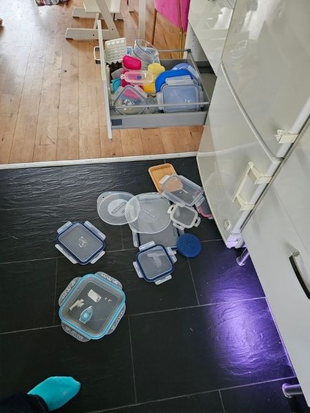 Schwarzer Küchenfußboden, eine offenstehende Schublade, und auf dem Boden Plastikdeckel von Aufbewahrungsdosen.