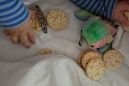 Kleine Babyhände, die aus einem gestreiften Schlafanzug hervorgucken, greifen nach kleinen Reiswaffeln, die auf dem Bett ausgekippt sind.