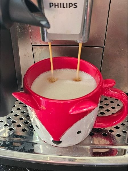 Kaffee läuft in eine rote Tasse mit Fuchsmotiv, in der sich schon geschäumte Milch befindet.