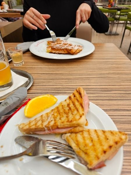 Zwei Teller auf einem Tisch. Über einem hantieren Hände mit Besteck an einer Waffel, auf dem anderen lieben zwei dreieckige gebackene Toast-Sandwiches.