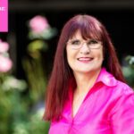 Die Romanautorin Petra Pflaum-Heinz lächelt vor einem natürlichen Hintergrund in die Kamera. Ihre Bluse ist pink, und im selben Pink befindet sich links oben im Bild ein Banner mit der Aufschrift: Petra Pflaum-Heinz: Warum ich nicht liefern muss.