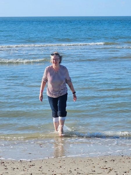 Silke Geissen kommt in einem rot-weiß-geringelten T-Shirt und einer hochgekrempelten Jeans ans Ufer, strahlend, durch die Wellen streifend.