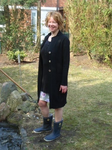 Silke Geissen steht lachend im eigenen Garten im Frühling, in langer schwarzer Strickjacke, geblümtem Rock und dunkelblauen Gummistiefeln, neben einem von Steinen umrandeten Teich.